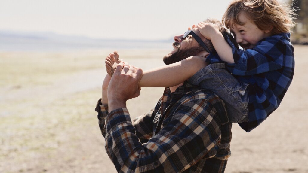 A photo of a smiling man with a child on his shoulders standing on a beach.