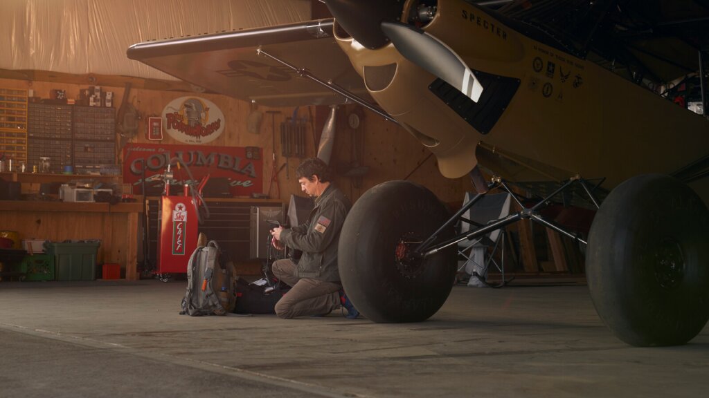 A pilot organizing his gear on the ground next to a brown colored bush plane.