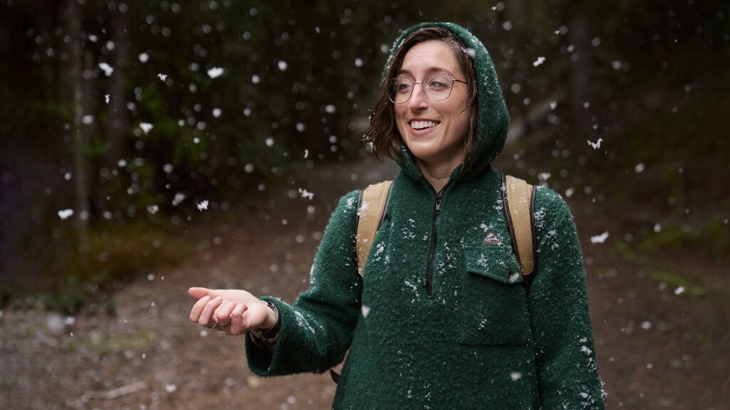 A smiling woman wearing a green, hooded jacket holding her hand out to catch large flakes of falling snow in the woods.