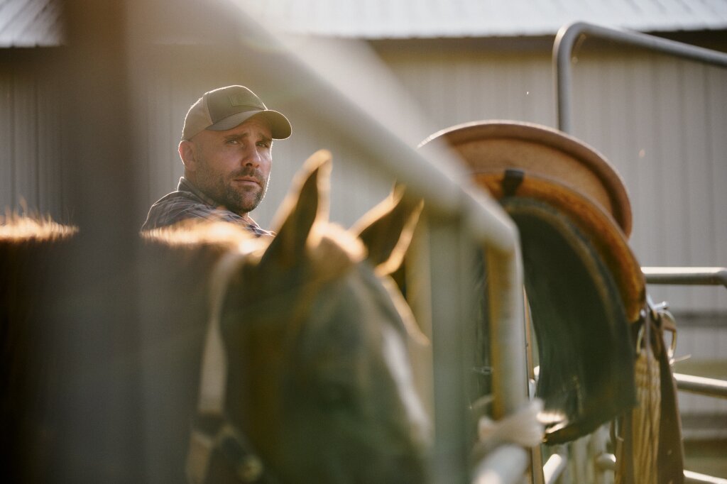 Ben Geisler standing at a fence, looking at the camera with a saddle on the fence and an out of focus horse in the foreground.