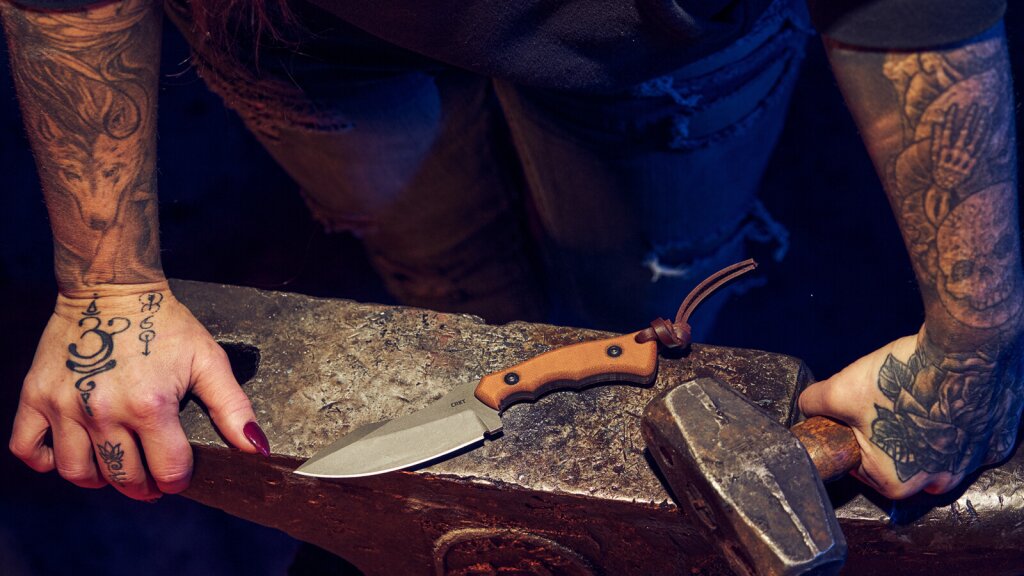 A photo of tattooed hands and arms resting on an anvil with a knife and hammer in the scene.