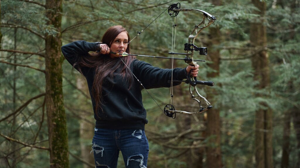 A photo of Kaila Cummings shooting an arrow in the woods.