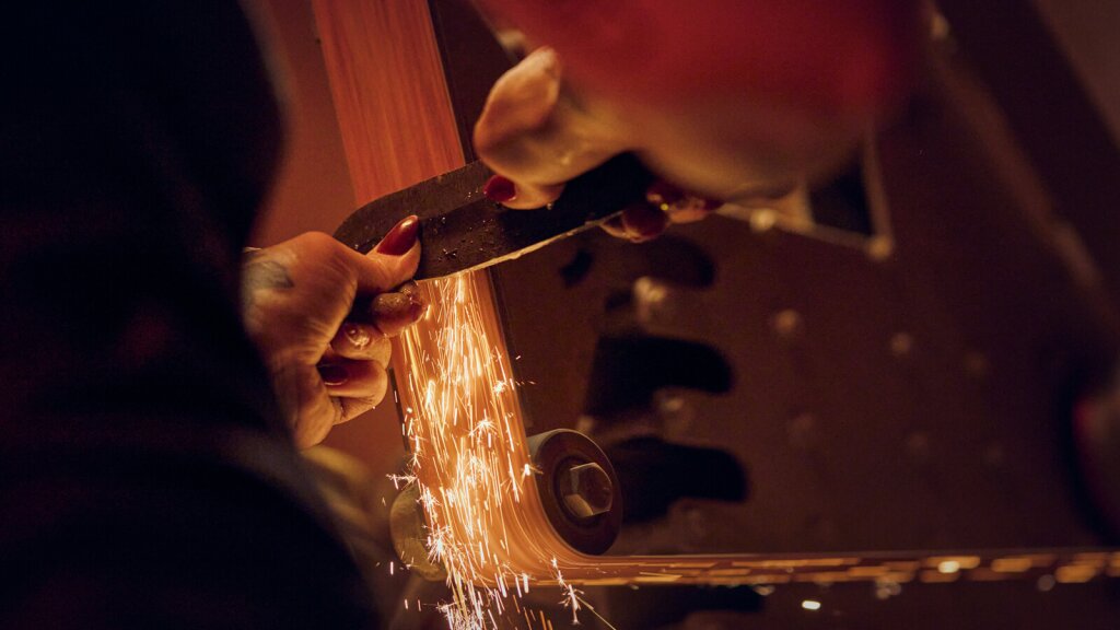 A close-up photo of a knife being sharpened on a belt sander with sparks flying.