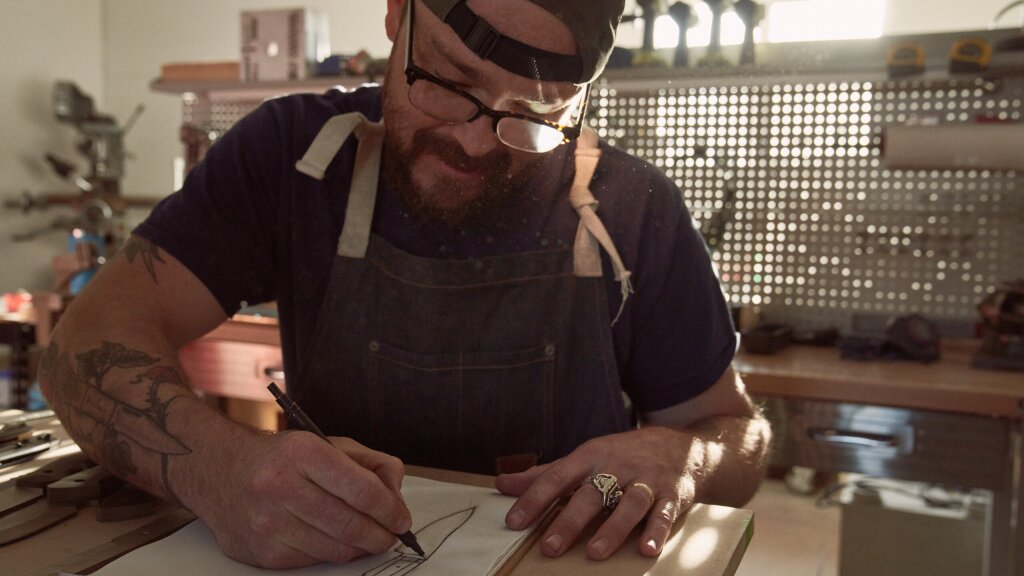 Lucas Burnley sitting at his workbench and sketching a design for a knife.