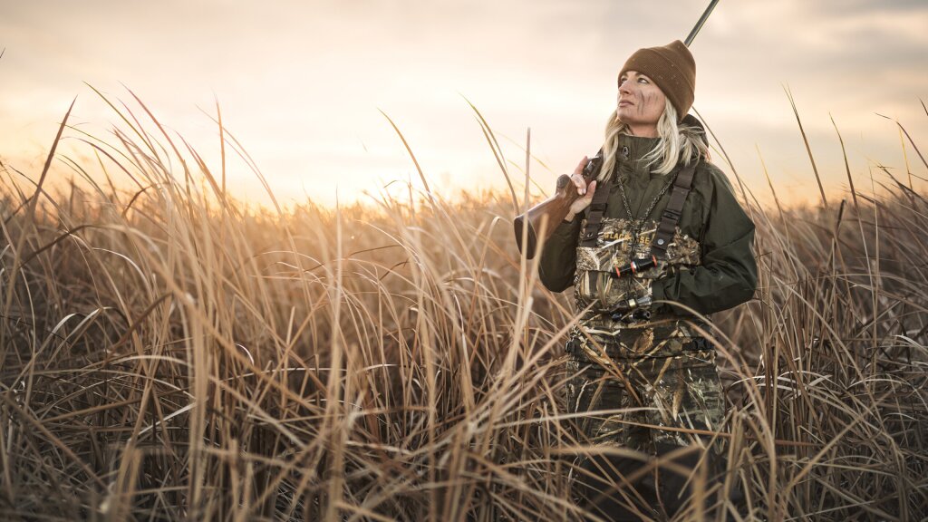 A female hunter standing in a field of tall, dry grass while holding a shotgun.