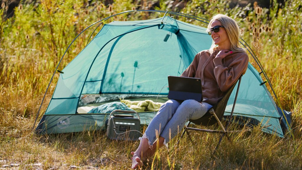 A photo of a smiling young woman working on her tablet in the backcountry next to her tent.